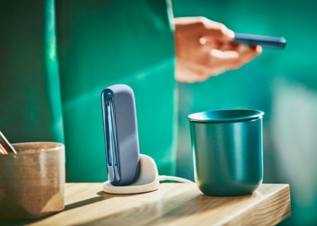 man holds an IQOS ILUMA mid azure blue device, with a holder and cup on the table.  man holds an IQOS ILUMA mid azure blue device, with a holder and cup on the table.