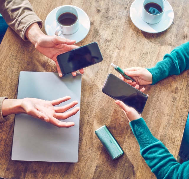 Two people holding mobile phones sit at a table; one also holds a jade blue IQOS ILUMA PRIME device.