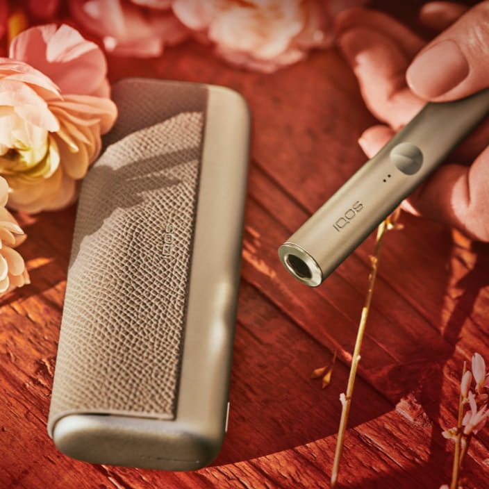 IQOS ILUMA prime device in gold khaki, set on a flower stall.